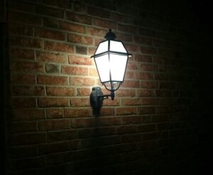 Außenleuchte mediterran mit Wandarm von Surya mediterrane Außenleuchte an einer Ziegelwand. Milchglas Scheiben. Lampe ist eingeschaltet. Viereckig mit 8 Lampengläsern.